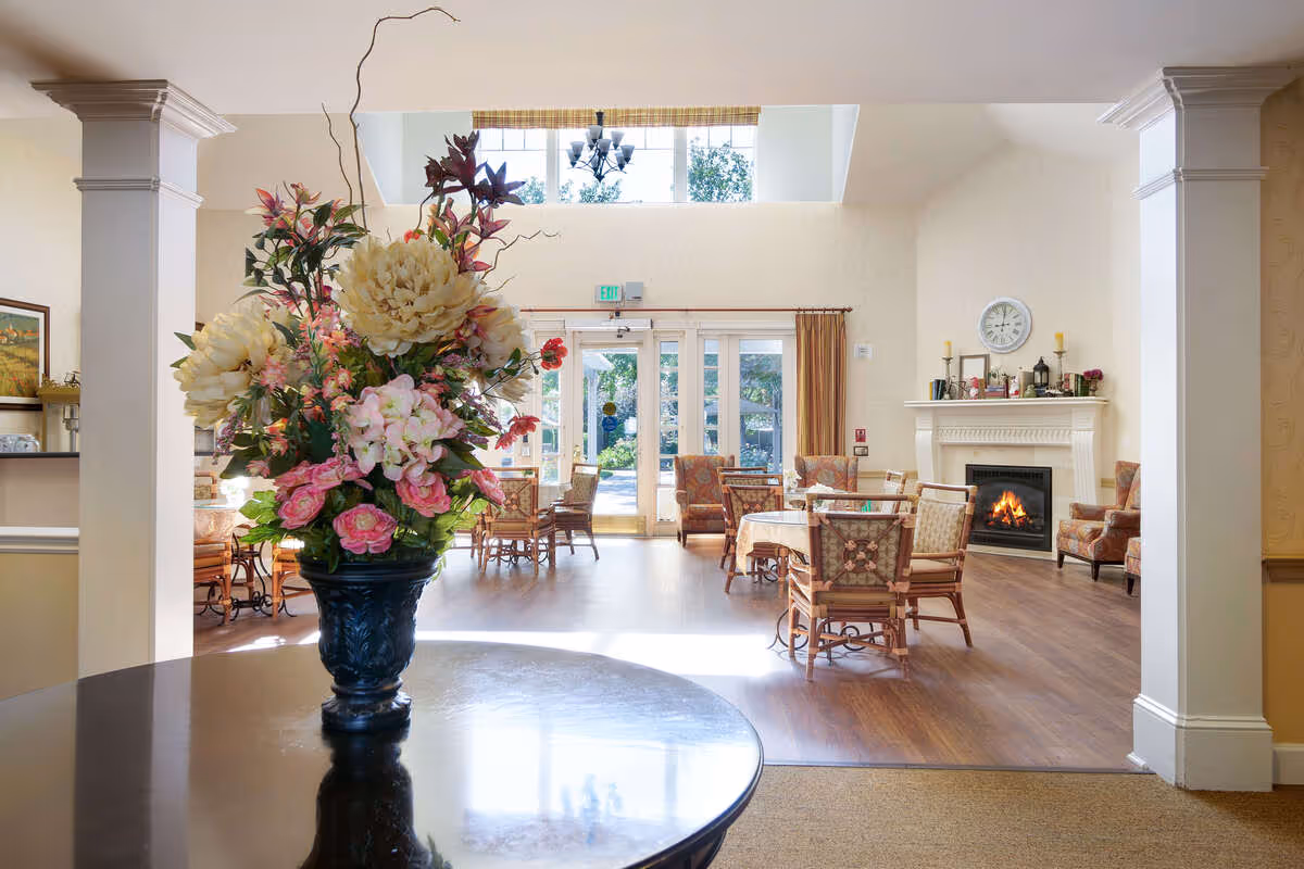 A bright and inviting senior living common area with a large floral arrangement in a black vase on a round table in the foreground. The room features wooden floors, multiple tables and chairs with floral upholstery, a lit fireplace with a mantel decorated with candles and small items, and large glass doors leading outside. Natural light fills the space through high windows above the doors.