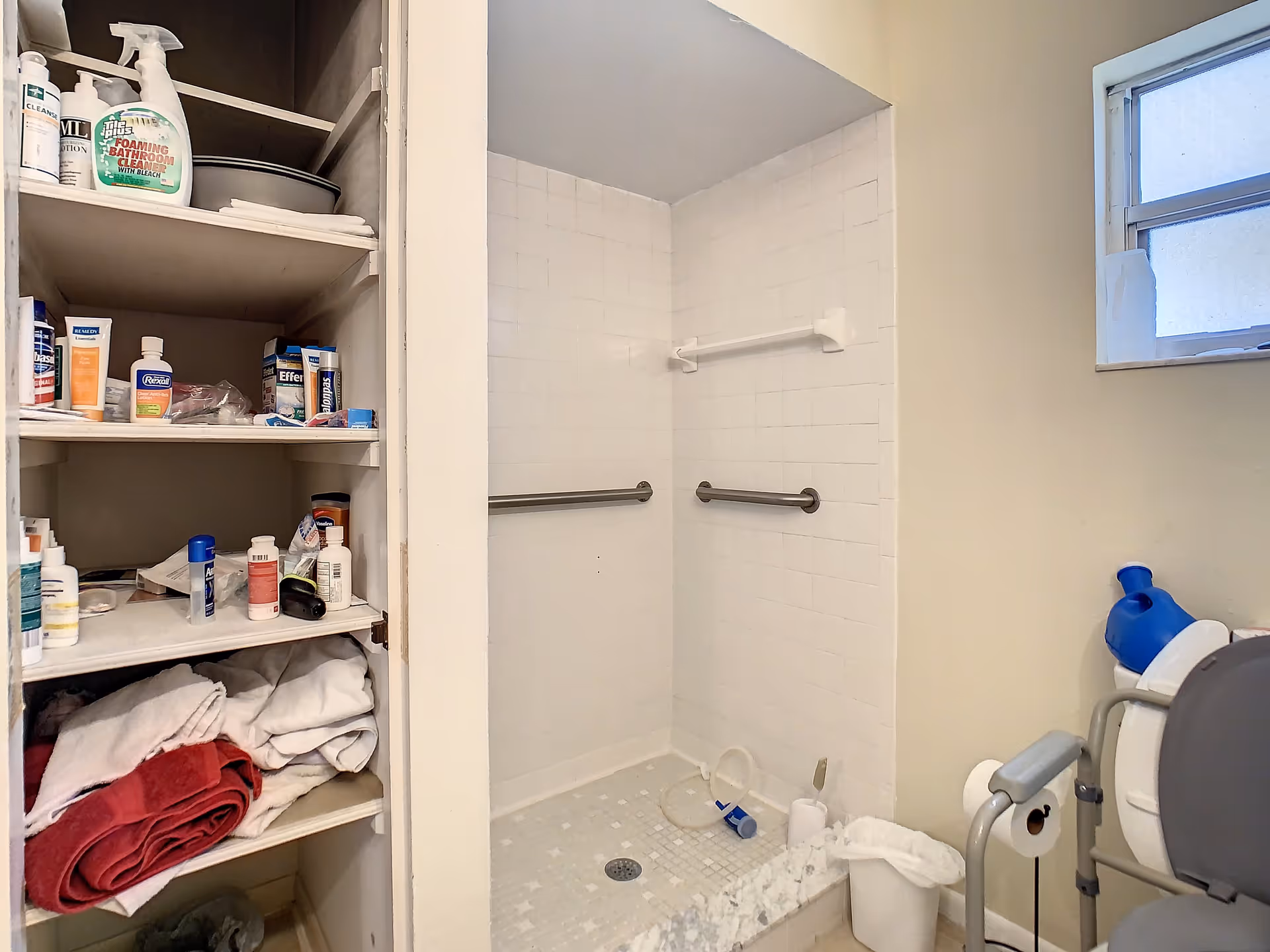 Bathroom with a walk-in tiled shower equipped with two grab bars. To the left, there is an open cabinet with shelves holding various toiletries and folded towels. On the right side, there is a toilet with a safety frame and a small window above it.