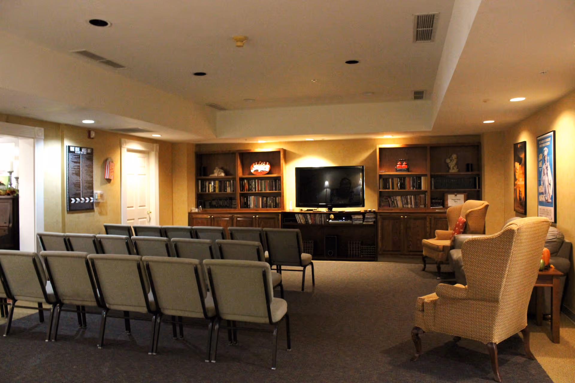 A cozy common area in a senior living facility with rows of beige chairs facing a large flat-screen TV mounted on a wooden cabinet with bookshelves. Two upholstered armchairs with patterned fabric are positioned to the right side near a small table. The walls are decorated with framed posters and the room is softly lit with recessed ceiling lights.