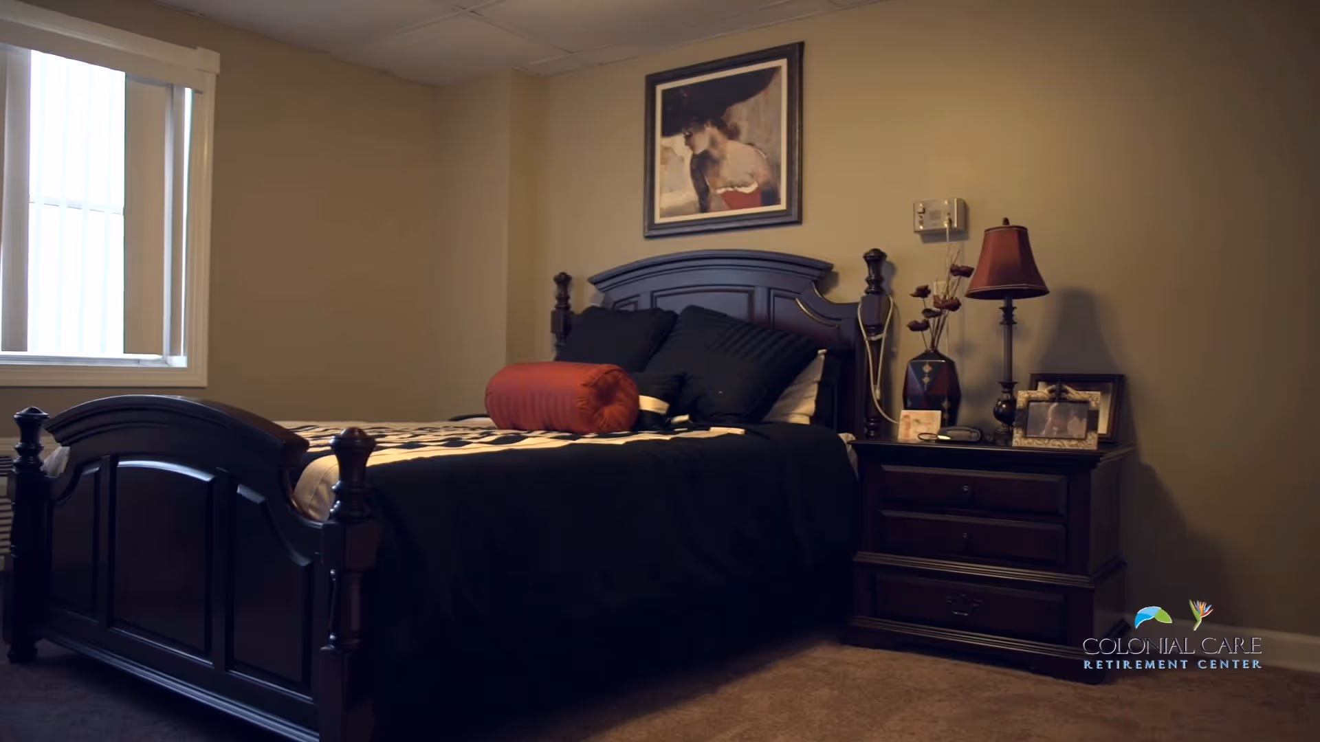 A bedroom with a dark wooden bed frame and matching nightstand. The bed is made with black and white bedding and has a red cylindrical pillow. On the nightstand are a lamp, a vase with decorative flowers, and framed photographs. A framed picture hangs on the beige wall above the bed. A window with vertical blinds is on the left side of the room.
