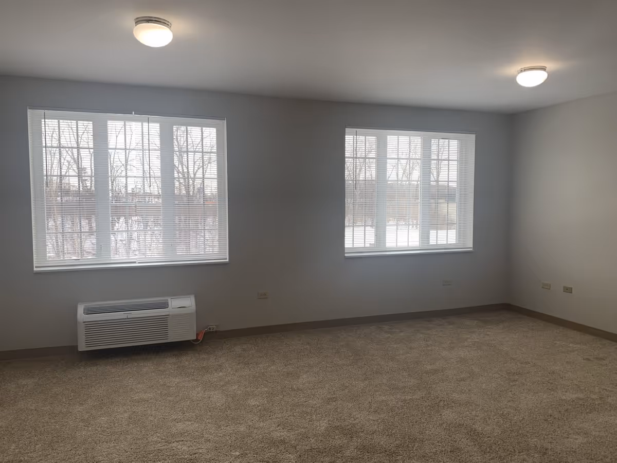 Empty room with beige carpet, two large windows with white blinds, white walls, two ceiling lights, and a wall-mounted air conditioning unit below one window.