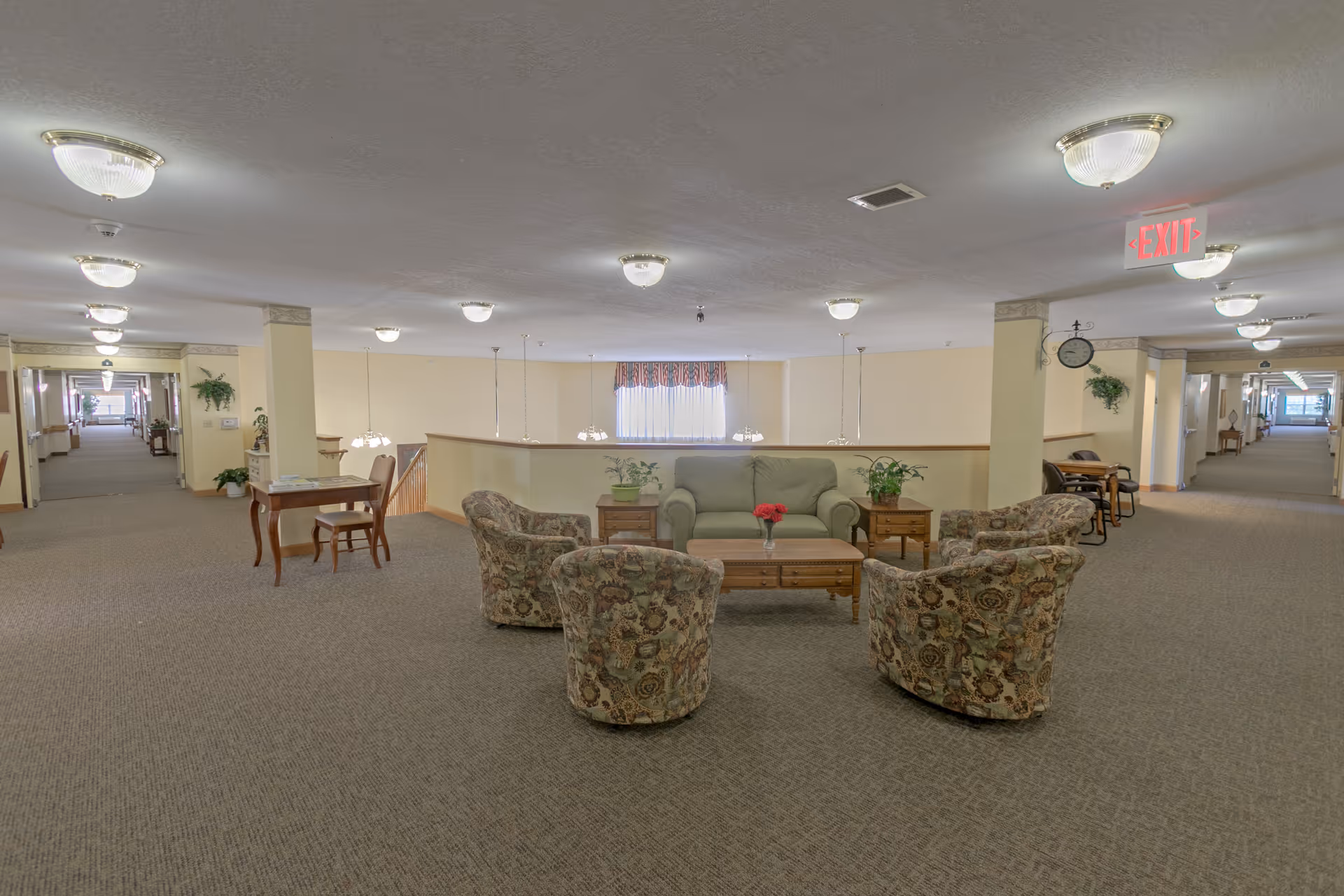 A spacious common area in a senior living facility featuring a green sofa, four patterned armchairs arranged around a wooden coffee table with a vase of red flowers, and side tables with plants. The area is carpeted and well-lit with ceiling lights. There are hallways extending on both sides with additional seating and tables, and an exit sign is visible on the ceiling.