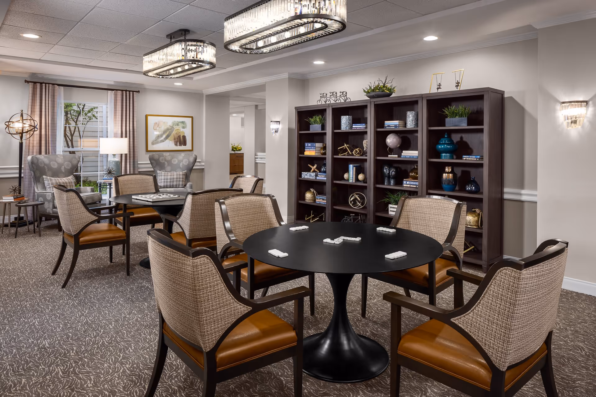 A well-lit senior living common area with a round black table surrounded by six cushioned chairs. The table has domino tiles placed on it. In the background, there are two armchairs with patterned upholstery near a window with curtains. A large dark wooden bookshelf filled with decorative items and books is against the wall. The room features modern ceiling lights and wall sconces, with neutral-colored walls and carpeted flooring.
