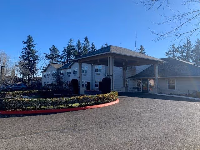 Front entrance of an assisted living facility with a covered porte-cochere, circular driveway, and landscaped shrubs under a clear blue sky.