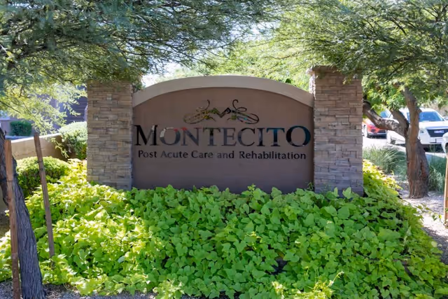 Stone and stucco sign for Montecito Post Acute Care and Rehabilitation surrounded by green leafy plants and trees with a few parked cars visible in the background.