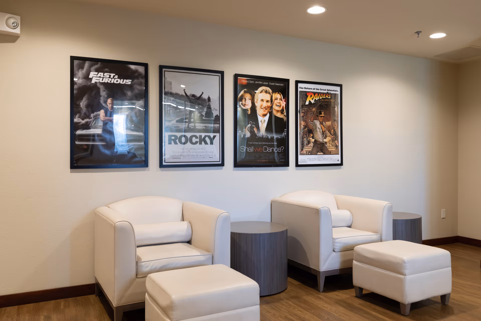 A cozy seating area with two white armchairs and matching ottomans, separated by two small round wooden tables. Four framed movie posters are hung on the beige wall above the chairs, featuring Fast & Furious, Rocky, Shall We Dance?, and Raiders of the Lost Ark. The floor is wooden, and the ceiling has recessed lighting.