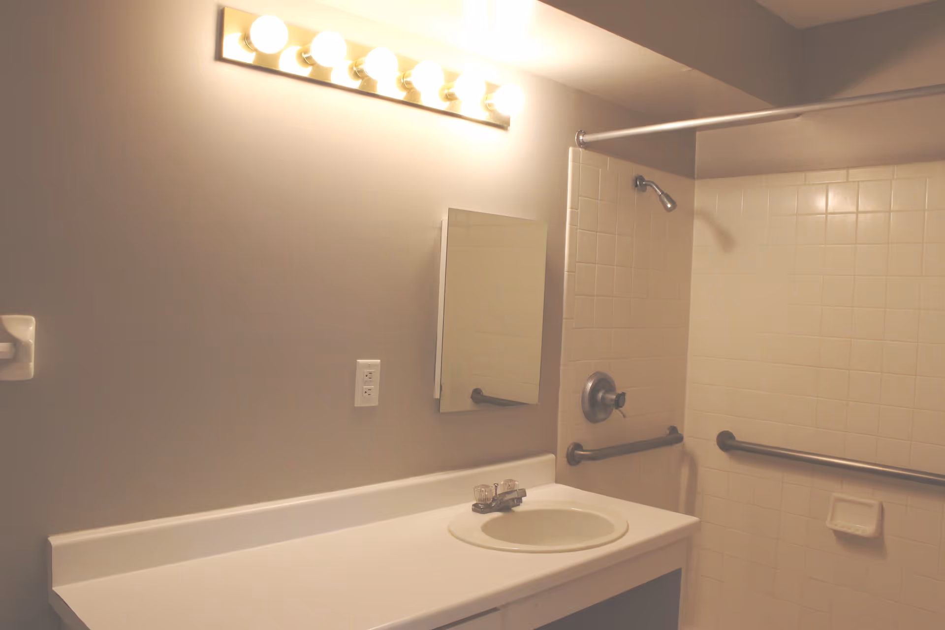 Bathroom interior with a white countertop and sink, a wall-mounted mirror, a row of six illuminated light bulbs above the mirror, and a tiled shower area with grab bars and a showerhead.