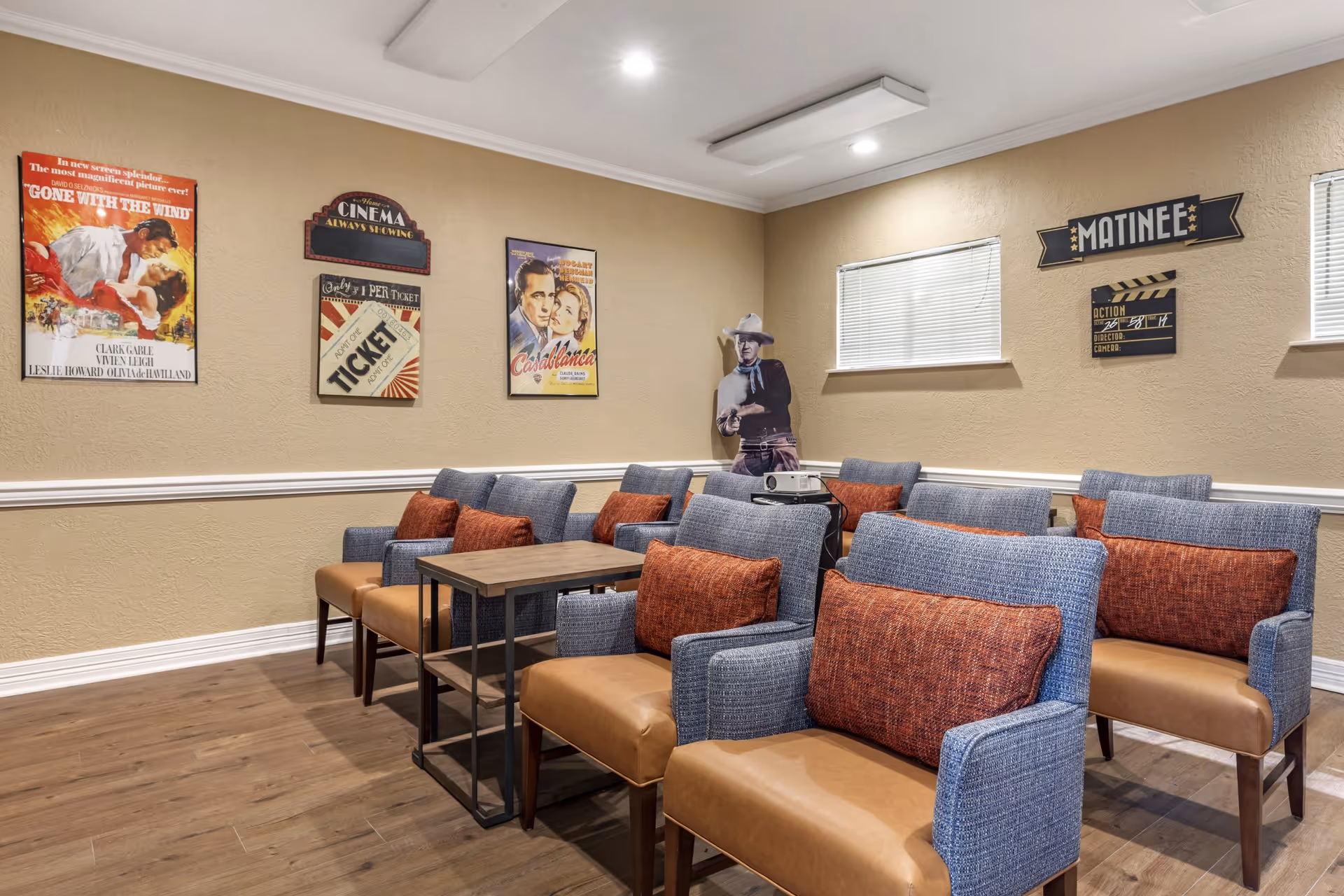 A small movie theater room with rows of blue and brown cushioned chairs, each with an orange pillow. The walls are beige with movie-themed decorations including vintage movie posters, a 'Matinee' sign, and a cardboard cutout of a cowboy. A projector is placed on a small table in the middle of the room.