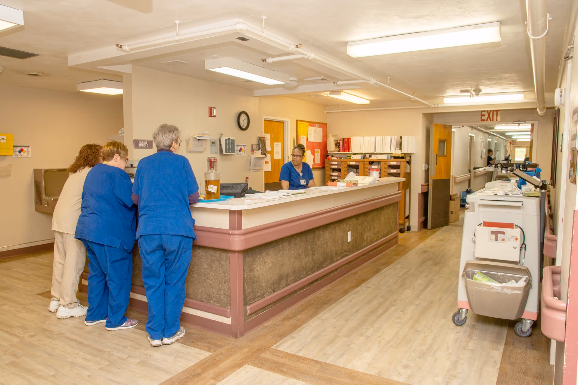 A nursing station inside a healthcare facility with three staff members in blue scrubs standing and talking to a seated staff member behind the counter. The area has light-colored walls, wood-patterned flooring, and medical carts along the hallway. An exit sign is visible at the end of the corridor.