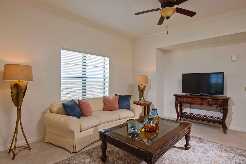 A cozy living room with a beige sofa adorned with blue and pink cushions, a wooden coffee table with decorative glass vases, two matching floor lamps with beige shades, a flat-screen TV on a wooden console, a ceiling fan, and a window with blinds letting in natural light.