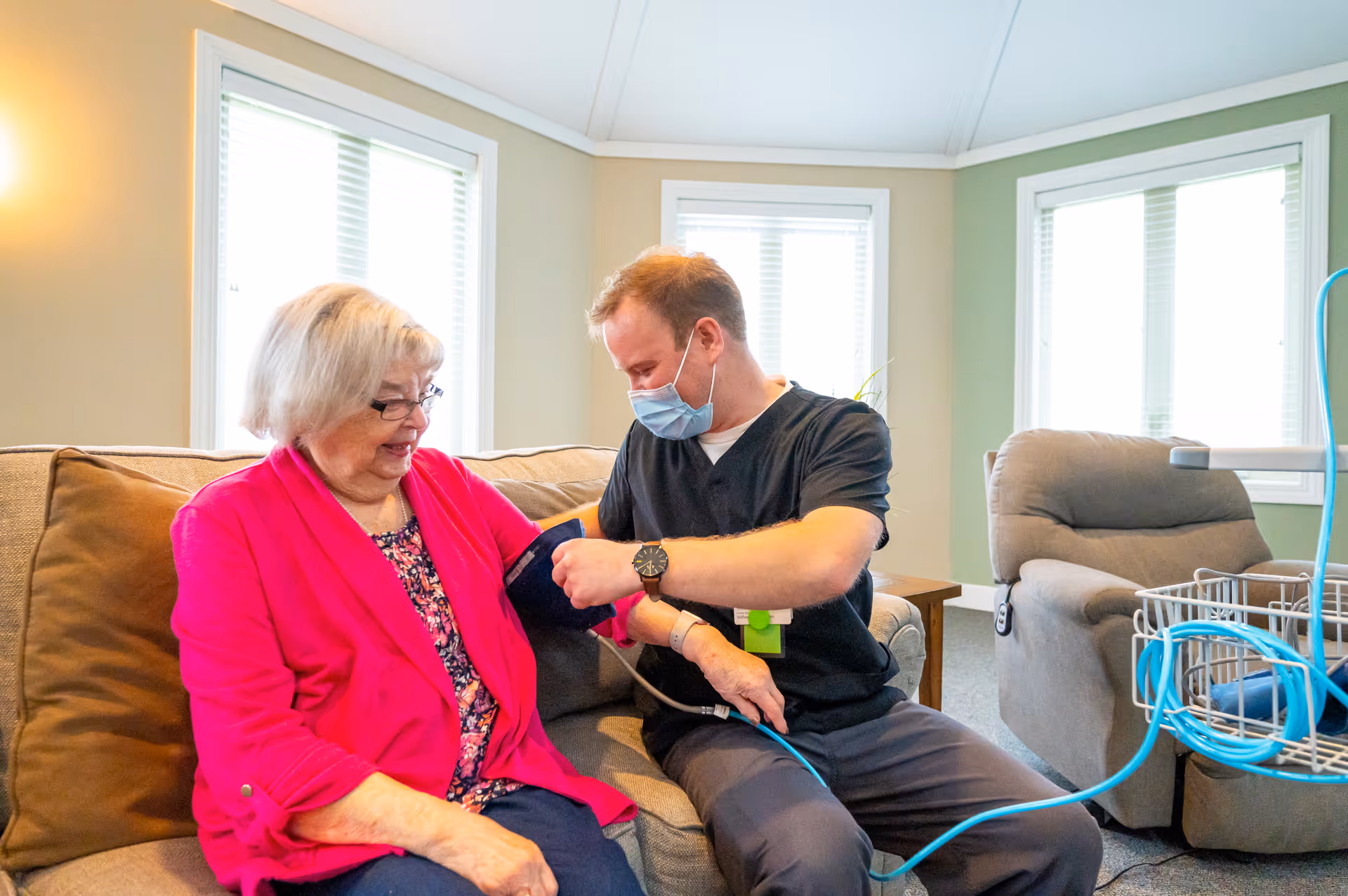 A healthcare worker wearing a mask is taking the blood pressure of an elderly woman sitting on a couch in a well-lit living room with large windows and comfortable seating.