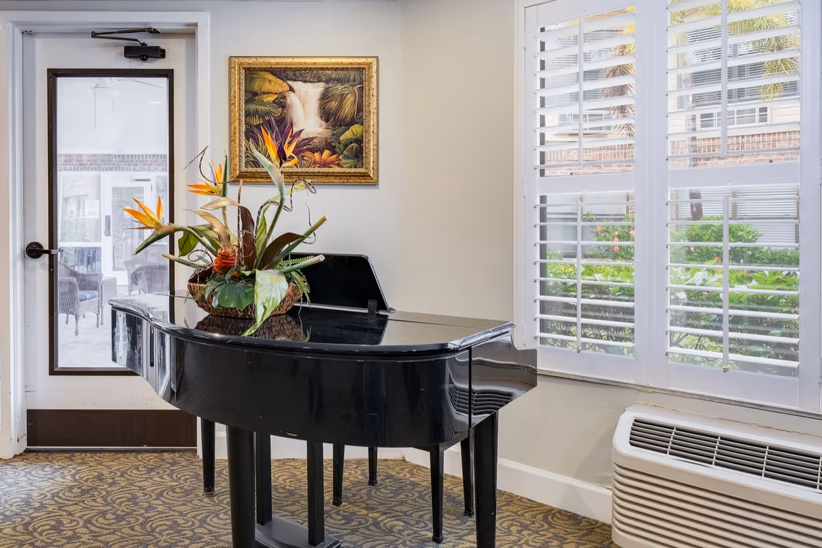 A black grand piano topped with a floral arrangement sits in a sunlit room next to shuttered windows and a glass door.