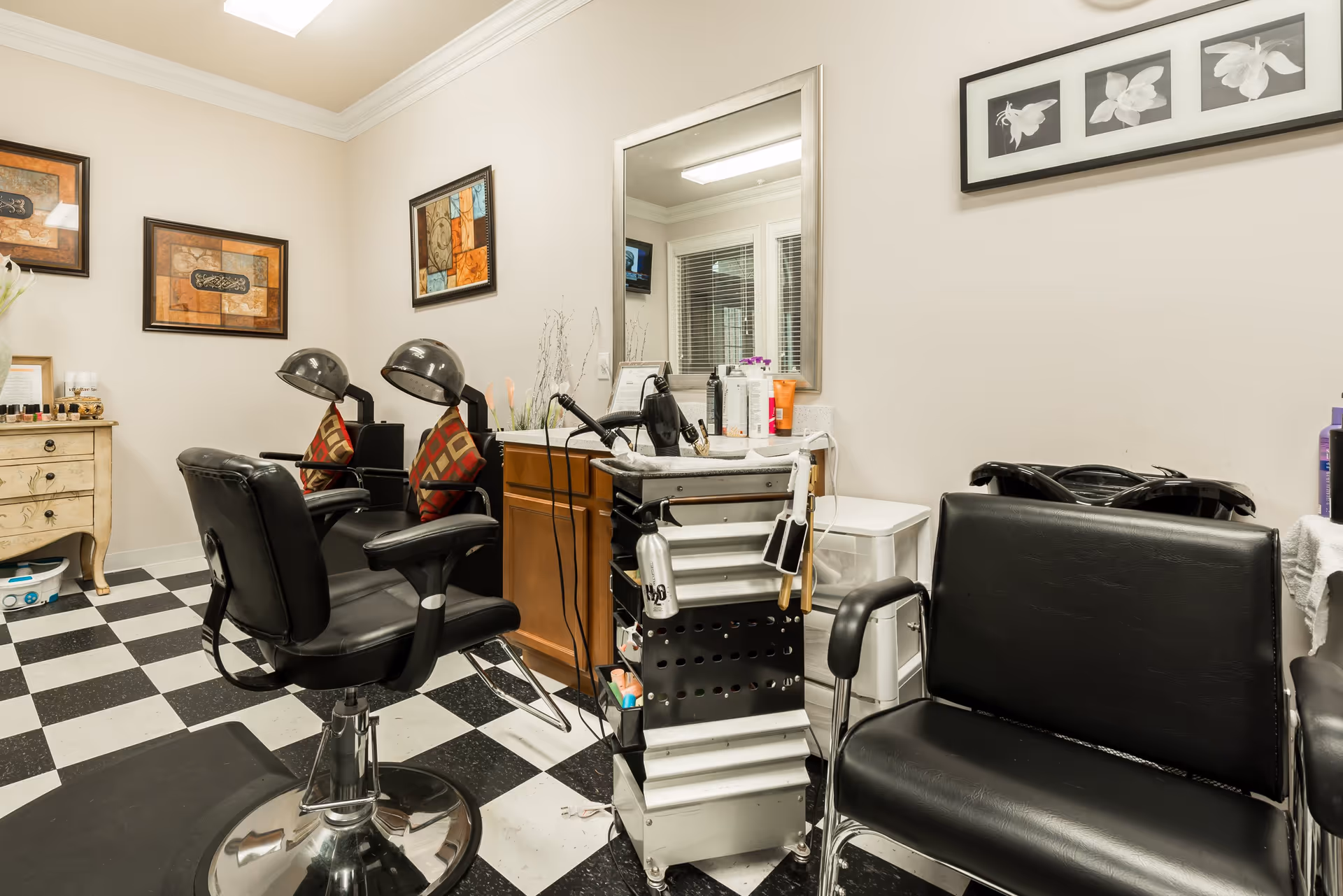 Interior view of a salon area with black and white checkered floor, two black salon chairs with hair dryers, a large mirror above a wooden cabinet, a rolling cart with hair styling tools, and framed artwork on the walls.