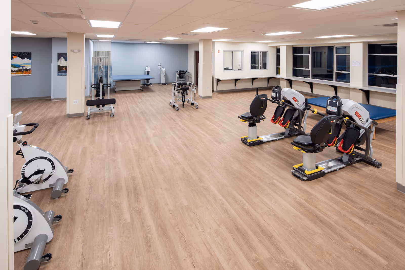 Large, well-lit rehabilitation gym with exercise machines, recumbent bikes, therapy tables, and wood-look flooring.