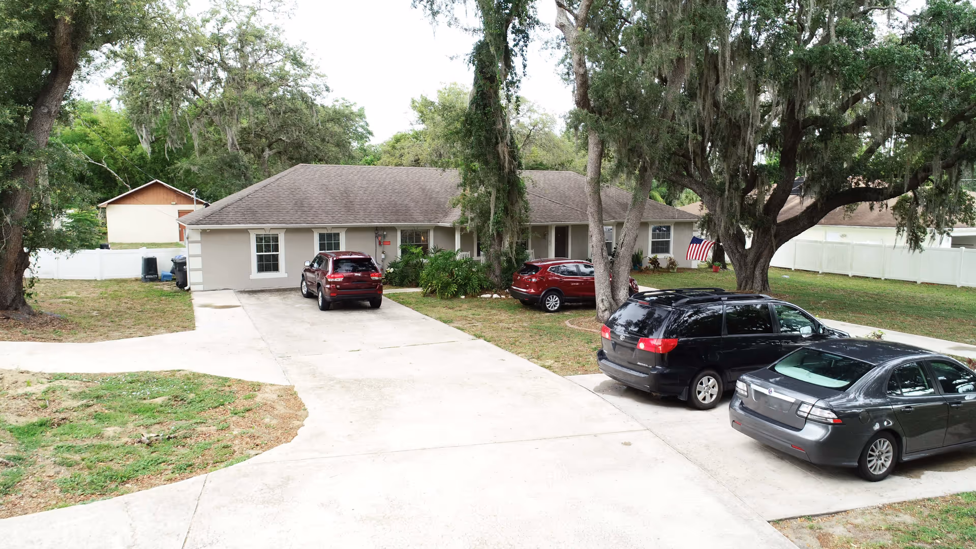 Exterior view of a single-story residential building with a gray roof and beige walls, surrounded by large trees and a white fence. There are four cars parked on the concrete driveway and lawn area, and an American flag is displayed near the entrance.