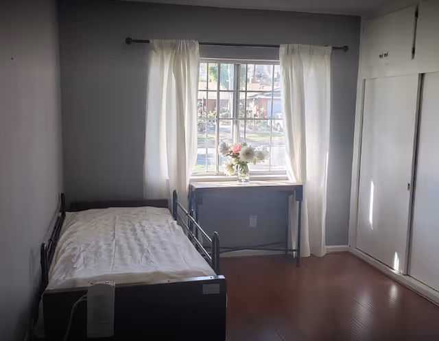 Small bedroom with a single bed, hardwood floor, a window with white curtains and a table holding a vase of flowers.