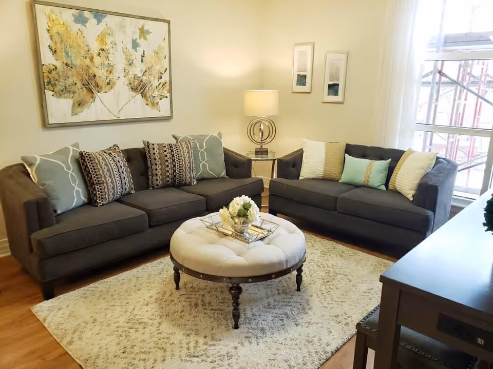 Bright furnished living room with two gray sofas, a round tufted ottoman coffee table, decorative pillows, lamp, and wall art.