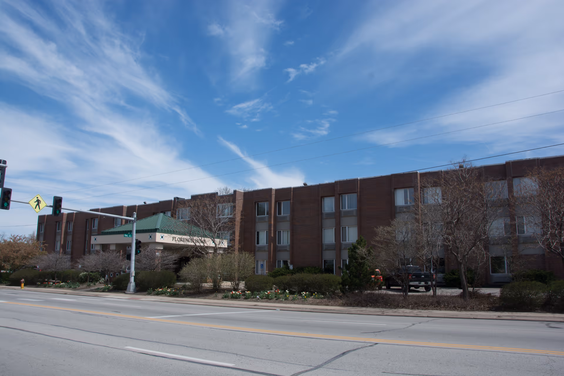 Exterior front view of a three-story brick senior healthcare building with a green-roofed entrance, landscaping, and a street in the foreground.