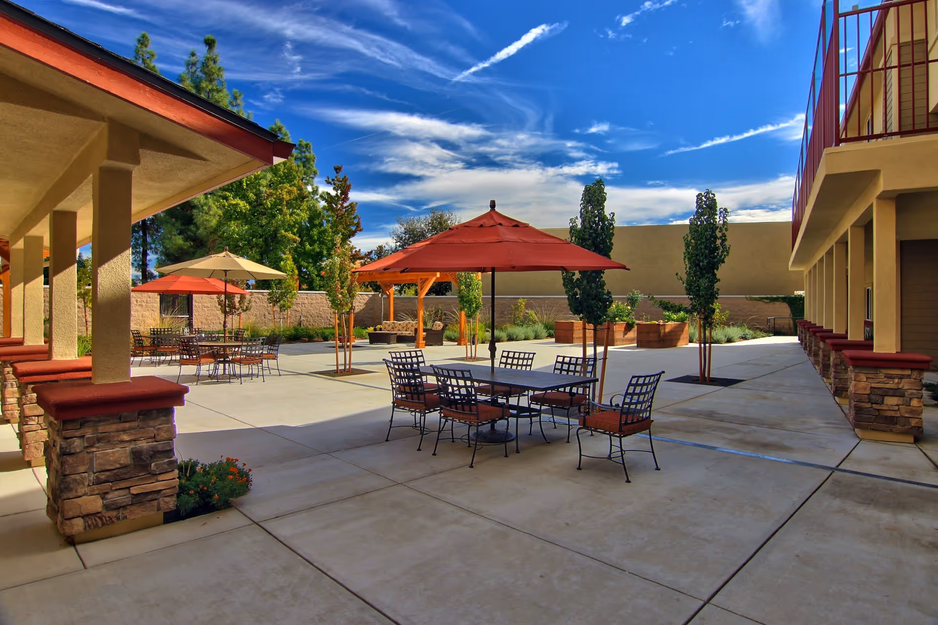 Outdoor patio area at Summerset Senior Living Rancho Cordova featuring several metal tables with chairs and large umbrellas providing shade. The patio is surrounded by a low wall and landscaped with small trees and plants. The sky is blue with some clouds, and the buildings have beige walls with red trim and stone accents.