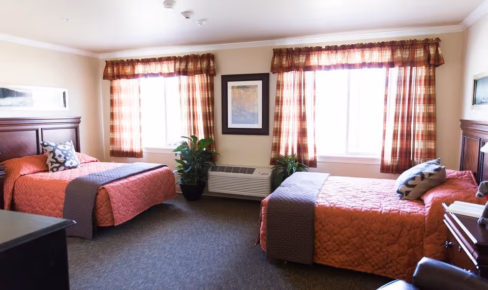 A bright bedroom with two twin beds, each with a reddish-orange quilt and a decorative pillow. The room has two windows with plaid curtains, a framed picture on the wall between the windows, two potted plants on the floor, and a heating/cooling unit beneath the windows. The furniture includes dark wooden headboards and a nightstand with a lamp.