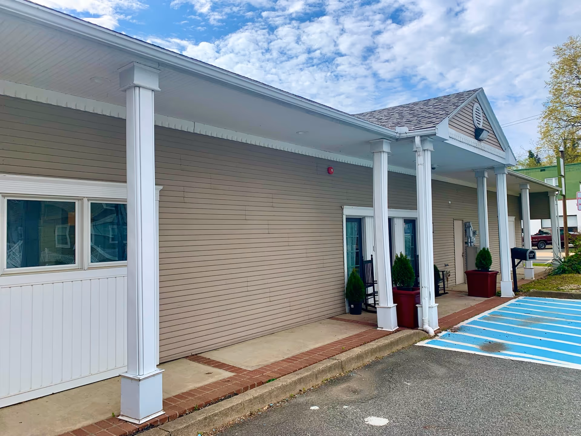 Exterior view of a single-story building with beige siding and white columns supporting a covered walkway. There are two large potted plants and two black rocking chairs near the entrance doors. The sky is partly cloudy and there is a blue and white striped handicap parking space in front of the building.