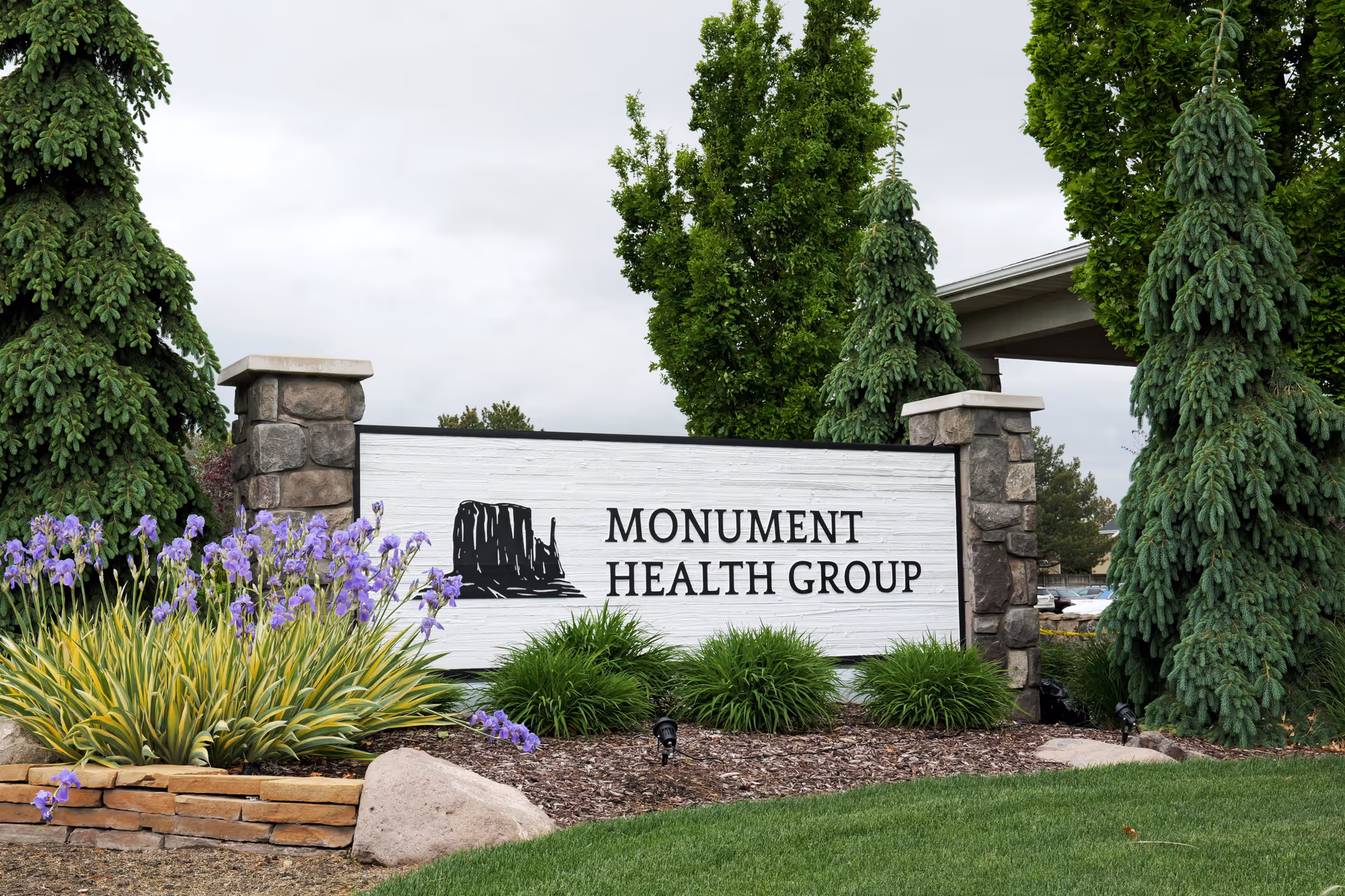 Outdoor view of a Monument Health Group sign surrounded by landscaping with green bushes, purple flowers, and tall evergreen trees under a cloudy sky.