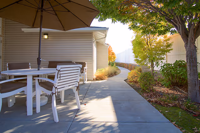 Outdoor patio area with a round white table and four chairs under a large beige umbrella. The patio is adjacent to a building with beige siding, and a concrete pathway curves alongside landscaped bushes and trees with green and autumn-colored leaves.