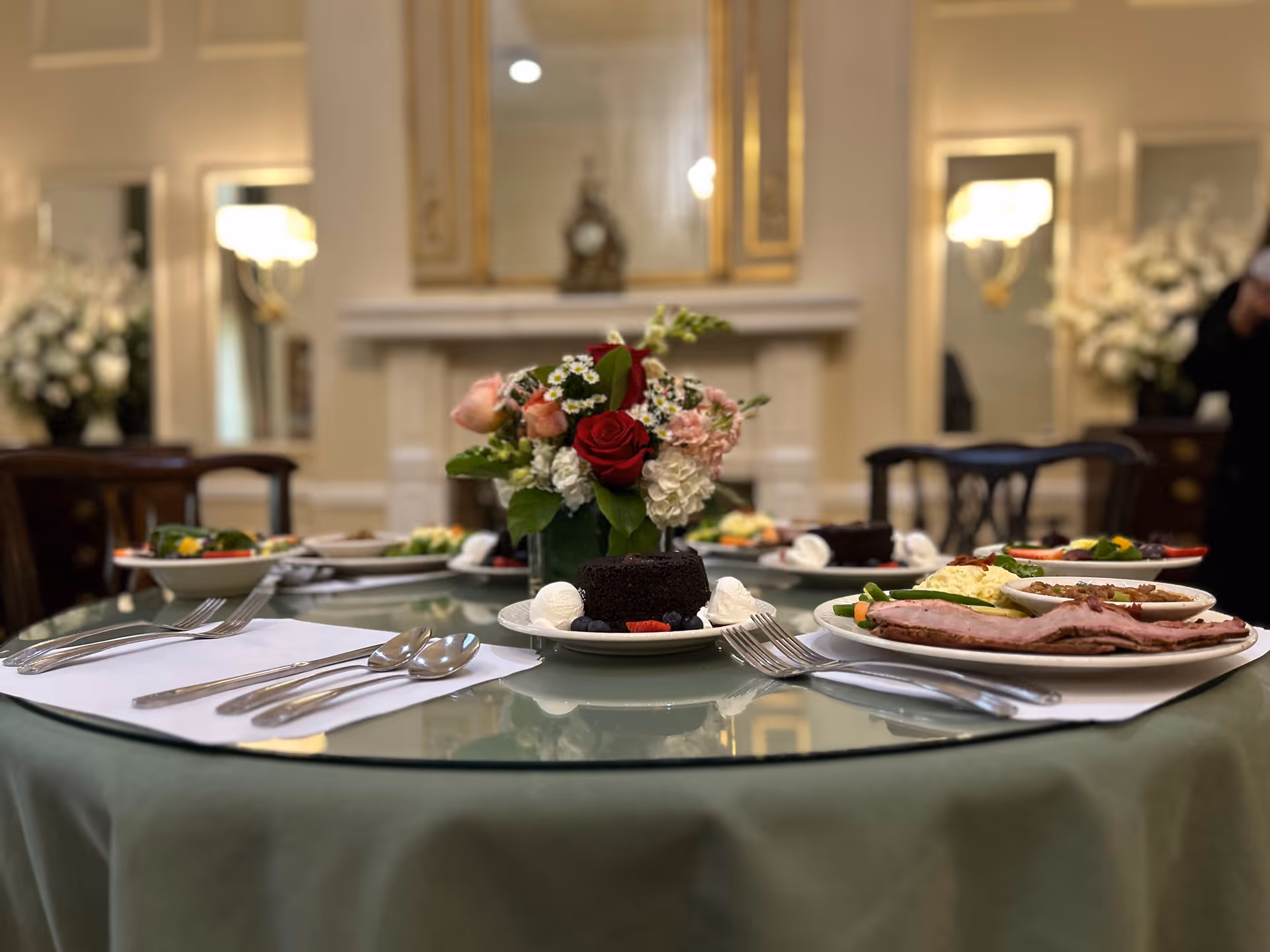 A dining table set with plates of food including slices of meat, salad, and dessert with whipped cream, arranged around a centerpiece of a floral bouquet with red and pink flowers. The background shows an elegant room with a fireplace, mirrors, and floral arrangements.