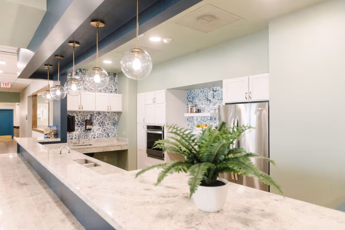 A modern kitchen area with a long marble countertop featuring a sink and a potted green fern plant. The kitchen has white cabinets, a stainless steel refrigerator, built-in oven, and blue patterned backsplash tiles. Five round pendant lights hang from the ceiling above the counter. The walls are painted light green and there is a hallway visible in the background.