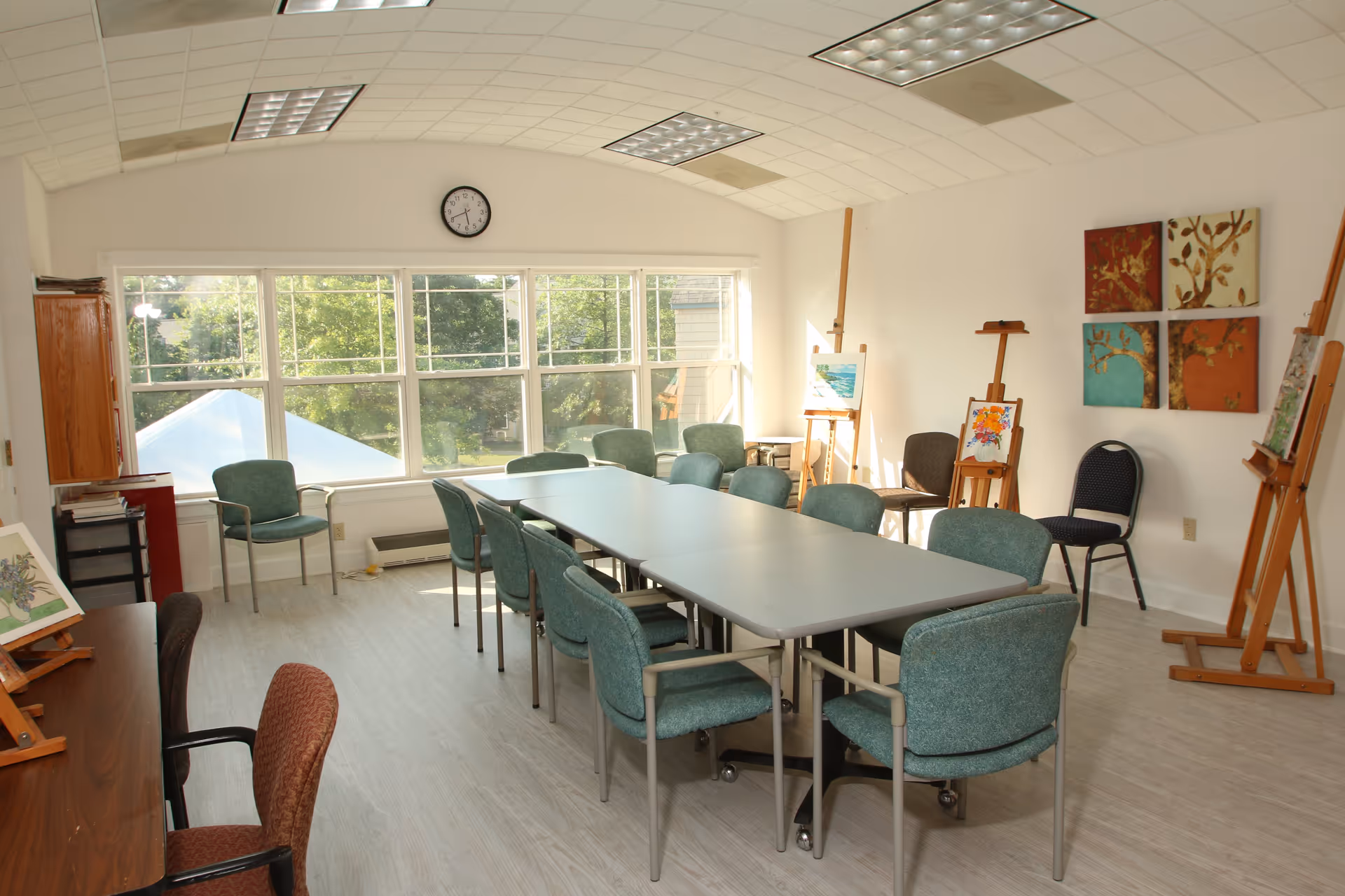 A bright art room with large windows letting in natural light. The room has a long rectangular table surrounded by green cushioned chairs. Several easels display colorful paintings, and there are additional chairs along the walls. The walls are white with four square paintings of trees in warm colors. A clock is mounted above the windows.