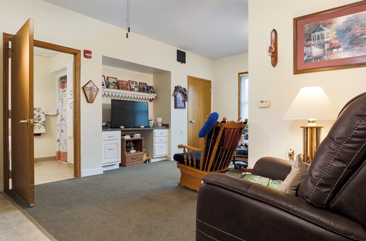 Interior view of a living room in an assisted living facility with a brown leather recliner, a wooden rocking chair with a blue cushion, a TV on a built-in white cabinet, and an open door leading to a bathroom with a shower curtain and towels.