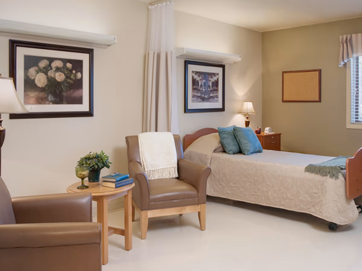 A cozy bedroom in a senior living facility with a single bed covered in a beige quilt, two blue pillows, and a green throw blanket. There are two brown leather armchairs with a small wooden table between them holding a plant and some books. The walls are decorated with framed artwork and a corkboard, and a window with blinds is visible on the right side.