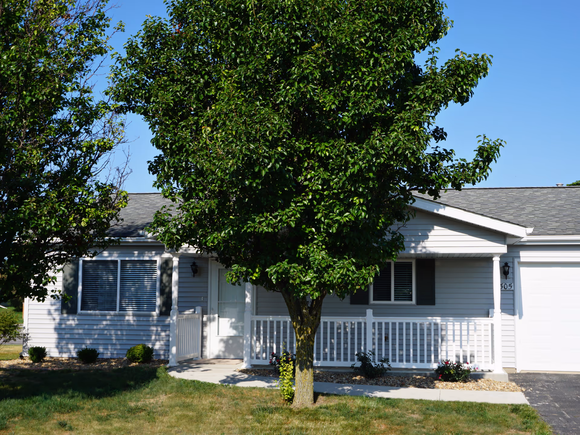 Front exterior view of a single-story white house with a small porch, white railing, and a tree in the front yard. The house has a gray shingled roof, two windows with shutters, and a driveway on the right side.