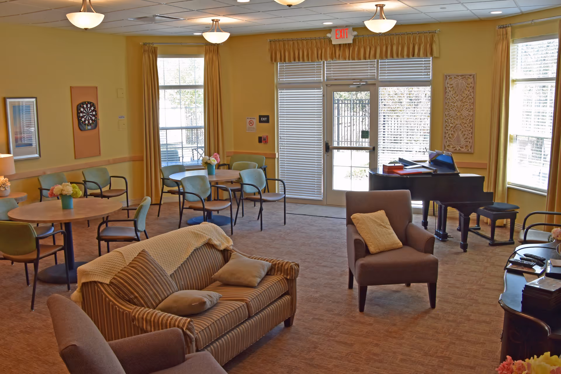 A cozy living room area with a striped sofa, armchair with a yellow pillow, round tables with chairs, a dartboard on the wall, and a black grand piano near large windows and a glass door with blinds. The walls are painted yellow and decorated with framed art and floral arrangements on the tables.