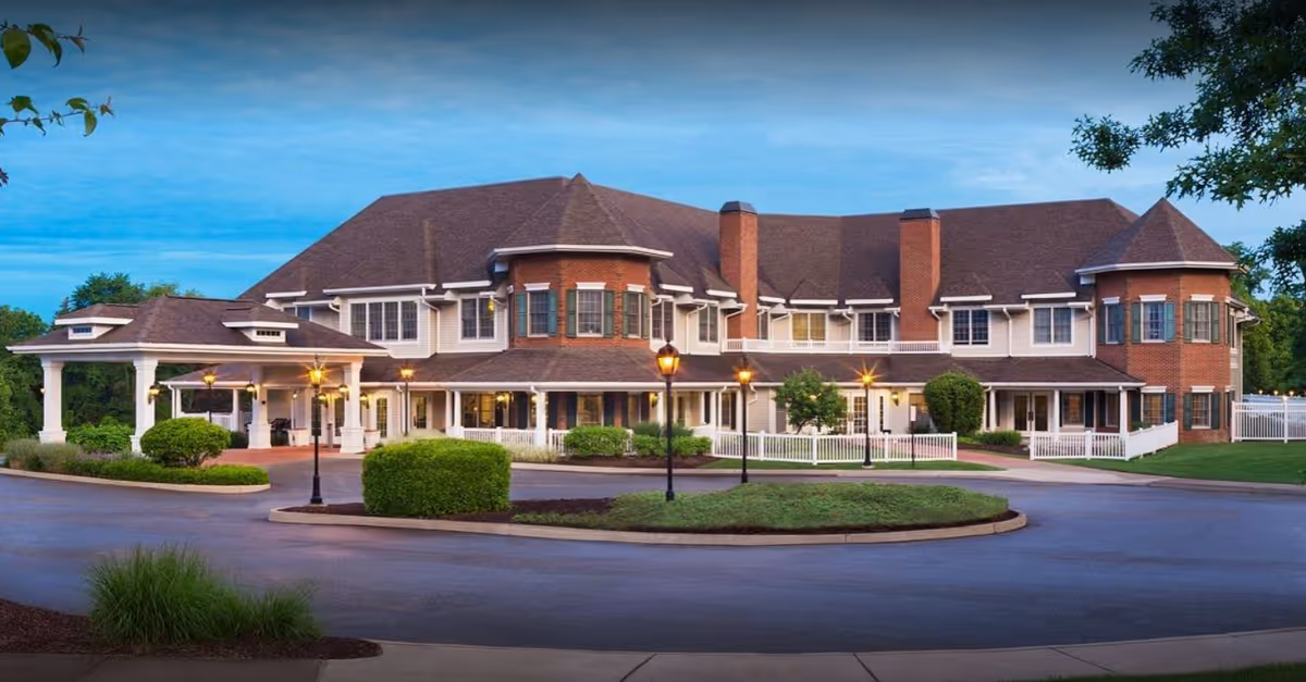 Exterior view of a large senior living facility building with a circular driveway, well-maintained landscaping, and multiple windows. The building features a combination of brick and siding with a peaked roof and a covered entrance area. Several lampposts are lit along the driveway.