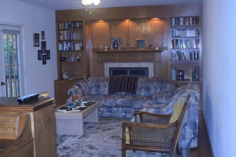 A cozy living room with a patterned sectional sofa, a wooden armchair with a yellow cushion, a coffee table with decorative items, and built-in wooden bookshelves surrounding a fireplace. There are framed photos on the mantel and a ceiling fan above. A sliding glass door is visible on the left side.