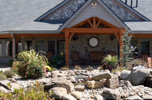 Outdoor view of a building entrance with a stone facade and wooden beams. There is a large clock mounted on the stone wall above a seating area with wooden chairs and a bench. In the foreground, there is a landscaped garden with rocks, plants, and a small water feature.
