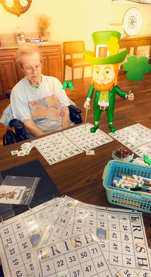 An elderly woman sitting in a wheelchair at a table playing a bingo game with cards labeled IRISH. A cartoon leprechaun holding a shamrock is digitally superimposed on the table. The room has wooden flooring and wooden furniture in the background.