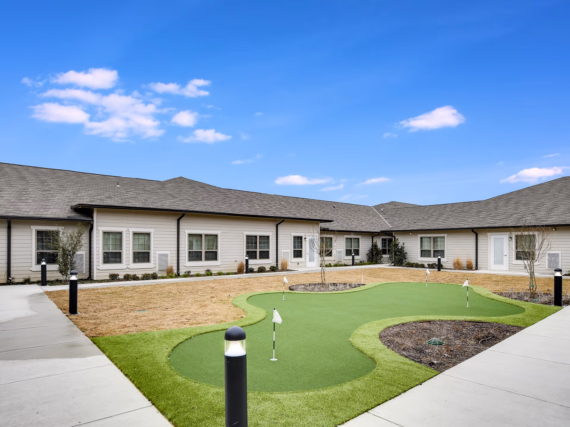 Outdoor courtyard area of The Grandview of Chisholm Trail featuring a small putting green with several golf holes and flags, surrounded by a concrete walkway and a single-story building with multiple windows and doors under a clear blue sky.