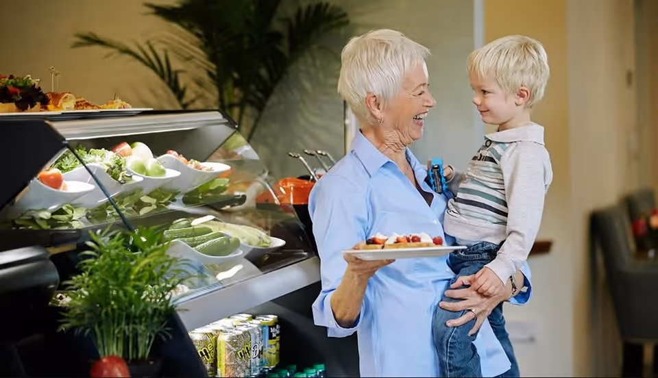 An elderly woman holding a young boy in her arms while smiling at each other in a dining area with a salad bar and beverages visible in the background.