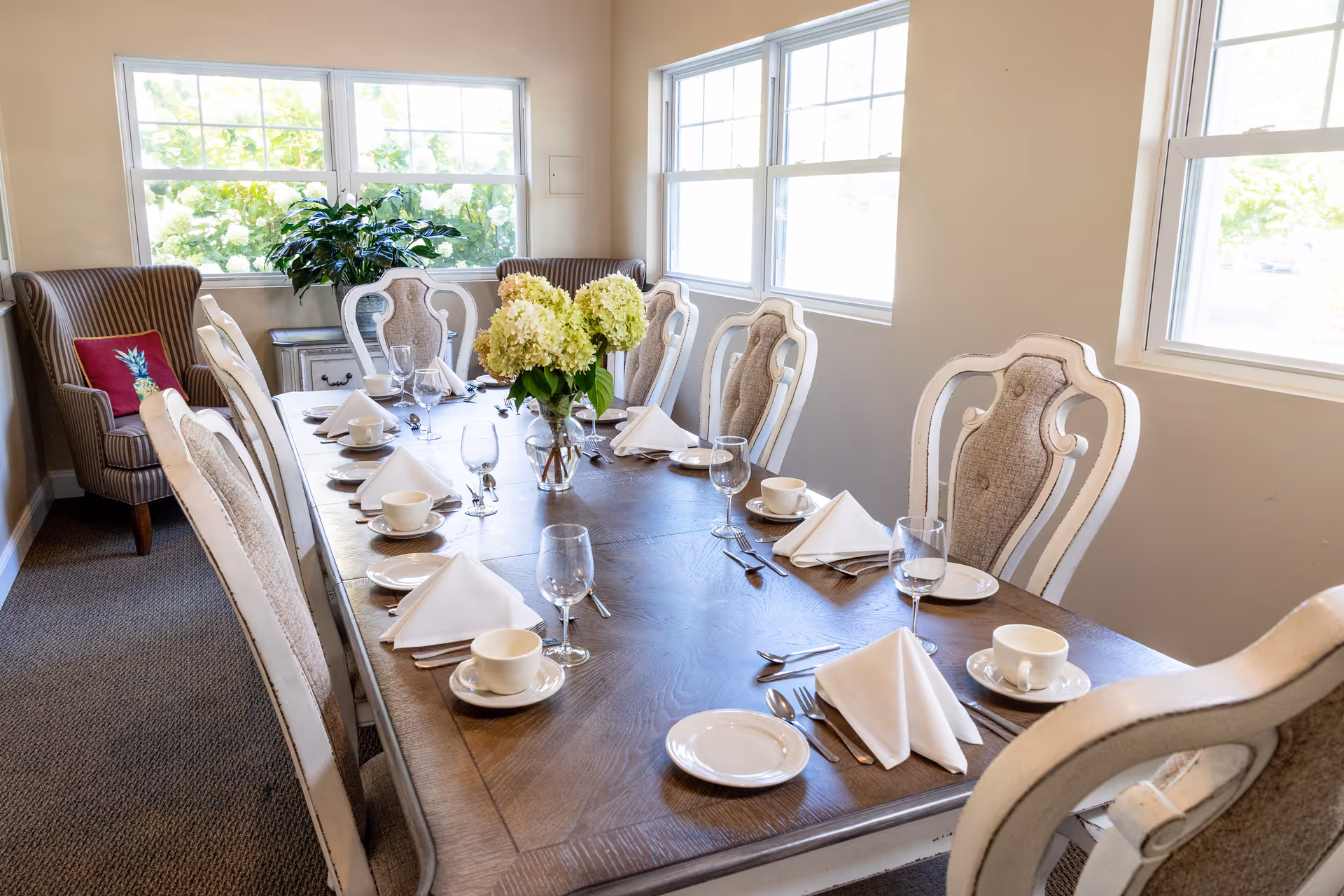 A dining room with a long wooden table set for eight people. Each place setting includes a white cup and saucer, a small plate, a folded white napkin, and silverware. There are two vases with light green hydrangea flowers in the center of the table. The room has beige walls, carpeted floor, and three large windows letting in natural light. A striped armchair with a red pillow featuring a pineapple design is positioned in the corner near the windows.