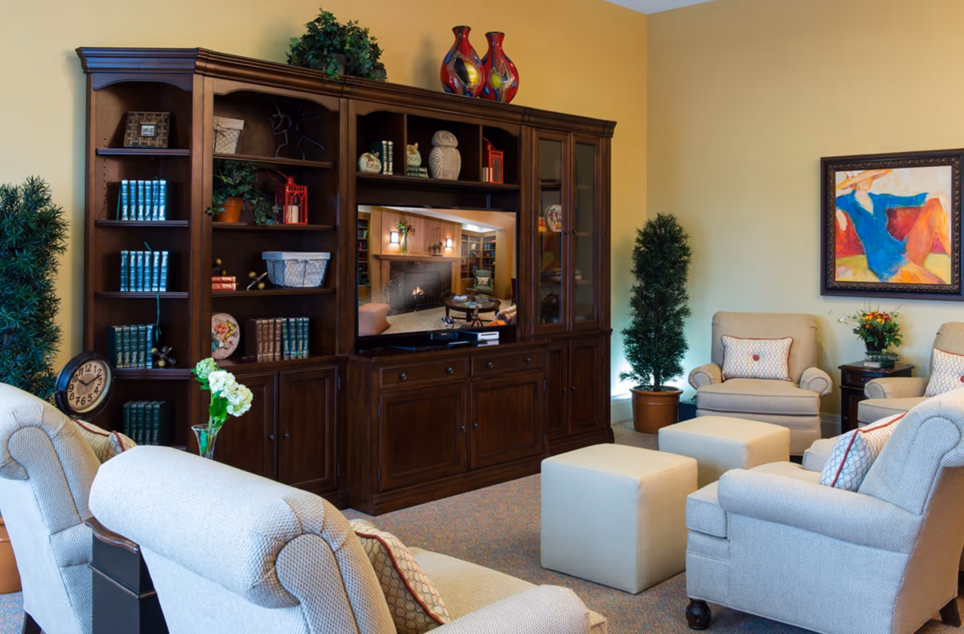 A cozy living room with beige upholstered armchairs and ottomans arranged around a dark wooden entertainment center with shelves holding books, decorative items, and plants. A flat-screen TV is mounted in the center of the unit, displaying an image of another room with a fireplace. The walls are painted light yellow, and there is a colorful framed painting on the right wall. Two potted plants flank the entertainment center.