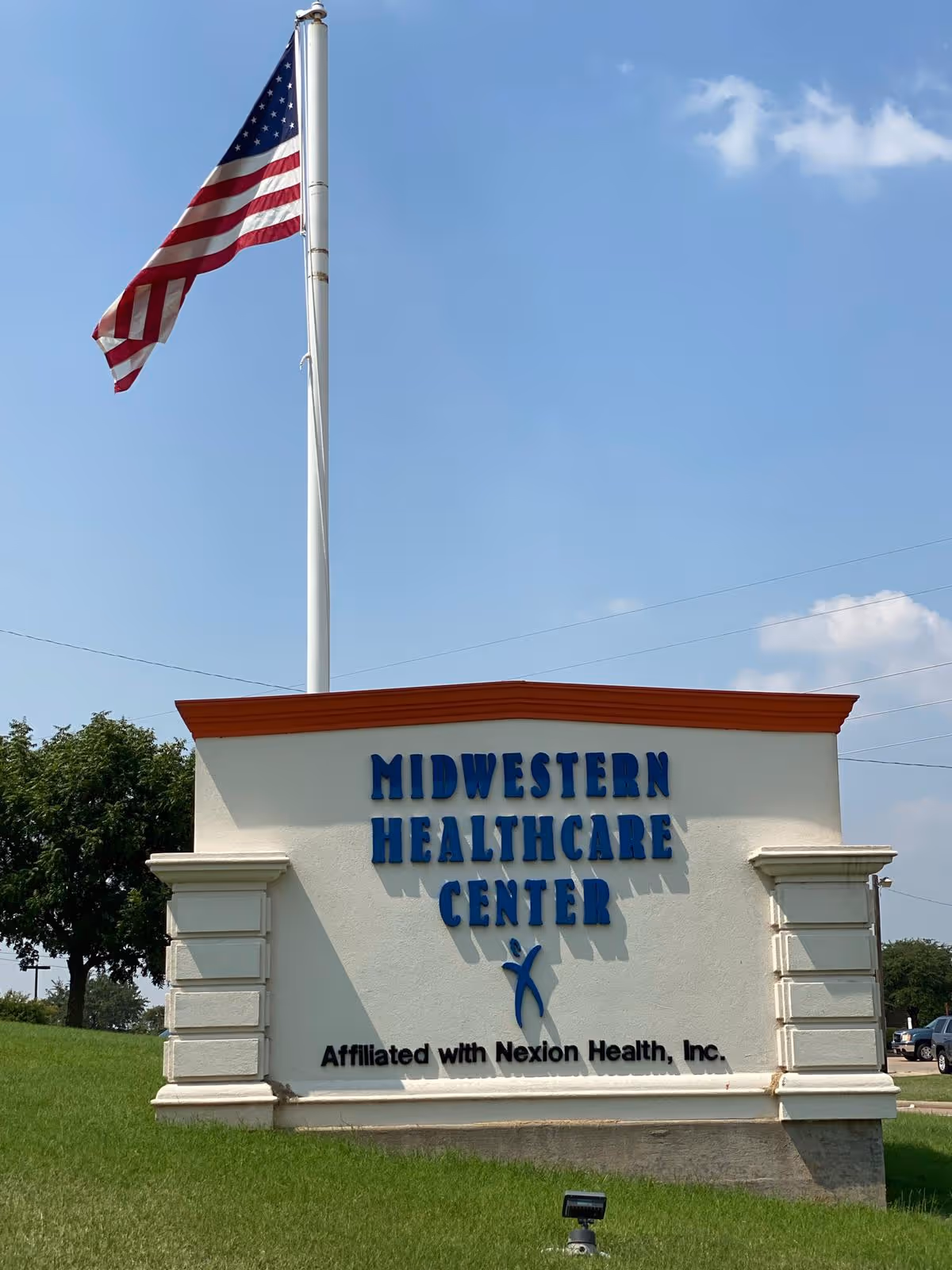 Outdoor sign for Midwestern Healthcare Center with an American flag on a flagpole behind it, set against a blue sky with some clouds and green grass in the foreground.