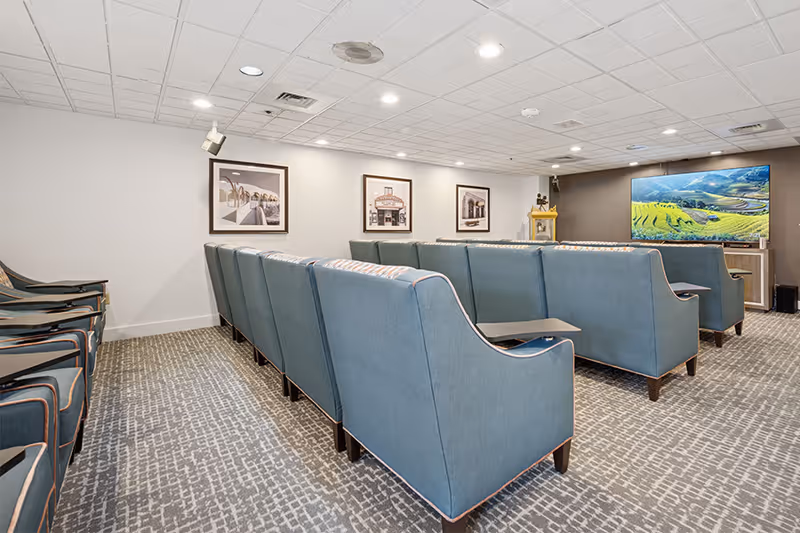 A small theater-style room with two rows of blue upholstered chairs facing a large flat-screen TV mounted on the wall. The room has a patterned carpet, white ceiling tiles with recessed lighting, and framed pictures on the walls.