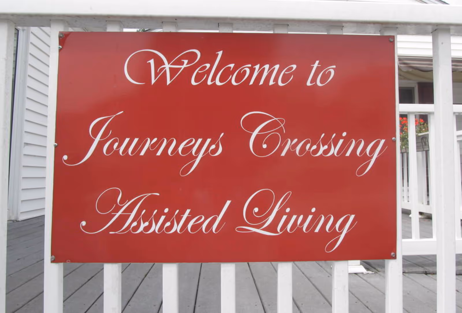 A red sign with white cursive text that reads 'Welcome to Journeys Crossing Assisted Living' mounted on a white wooden fence with a porch and part of a building visible in the background.