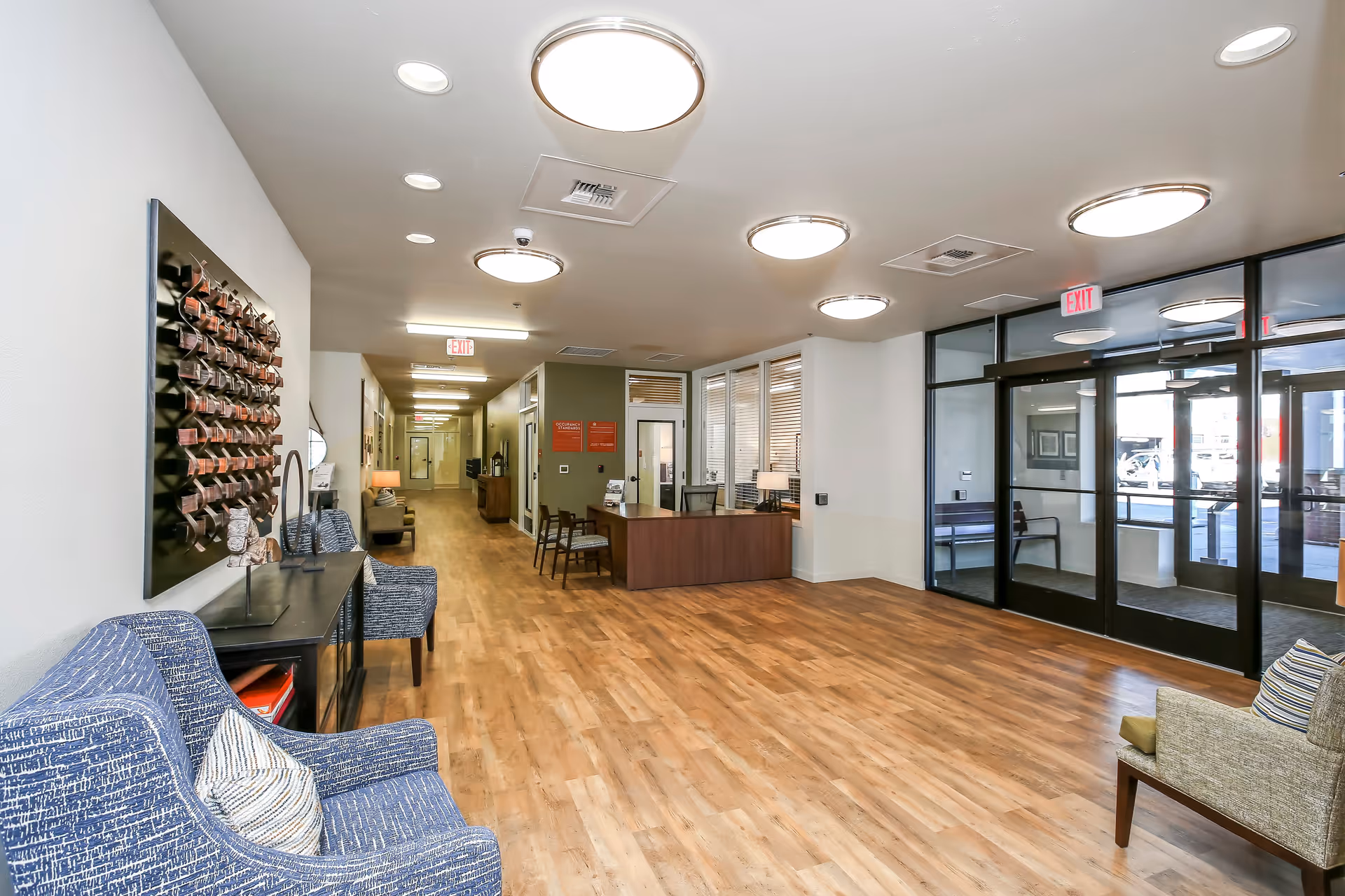 A spacious and well-lit lobby area with wooden flooring, blue and beige armchairs, a reception desk with chairs, and large glass entrance doors. The ceiling has multiple round light fixtures and there is a decorative wall piece on the left side.