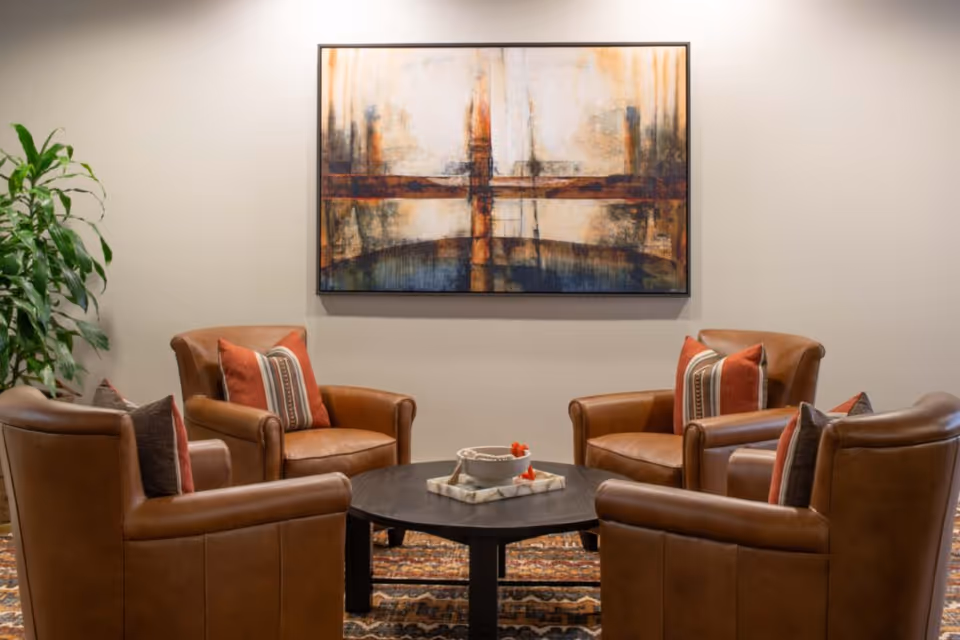 Four leather armchairs arranged around a round coffee table beneath abstract wall art in a cozy seating area.