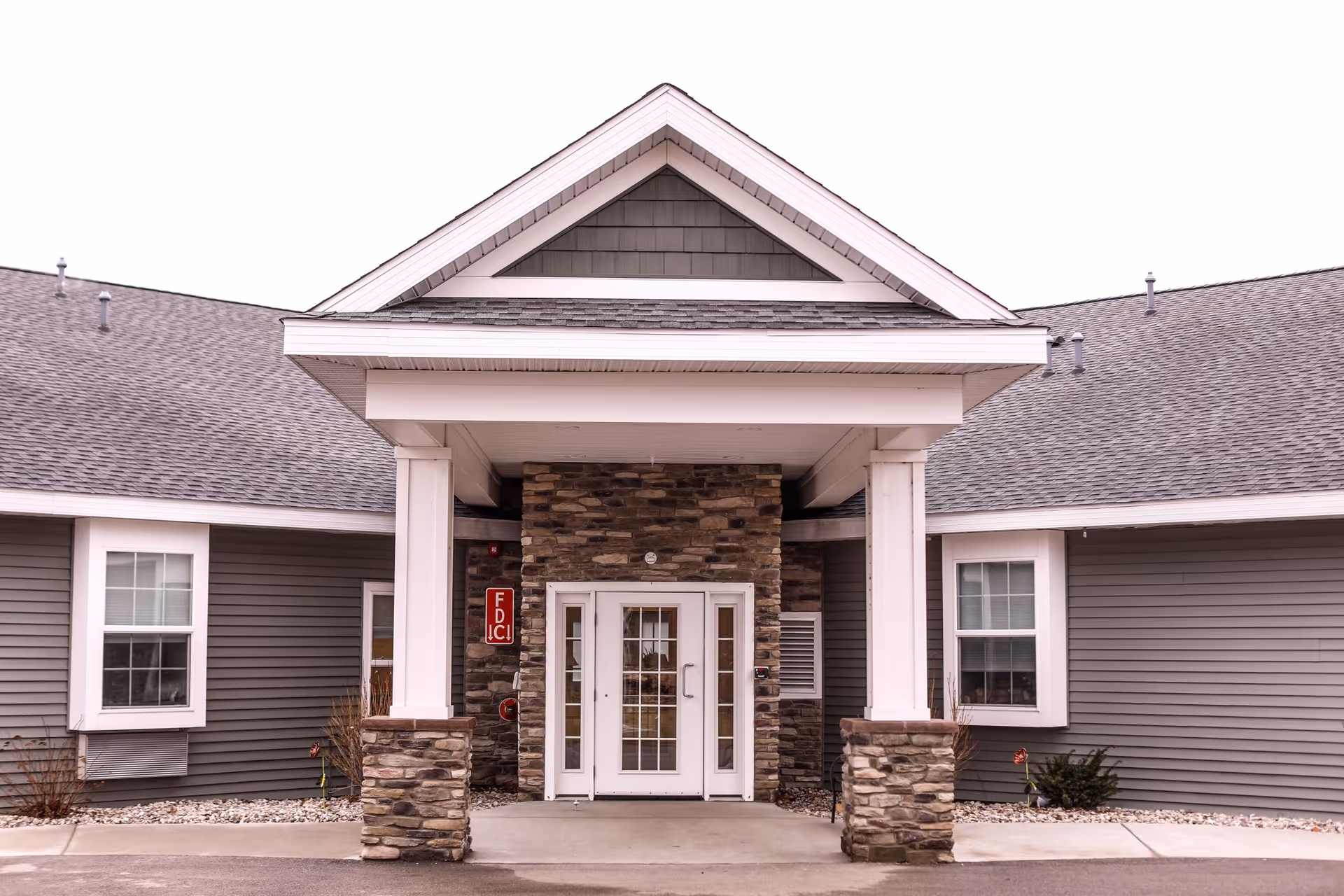 Front entrance of a building with a covered porch supported by white columns with stone bases. The building has gray siding, two windows on either side of the entrance, and a peaked roof above the porch.
