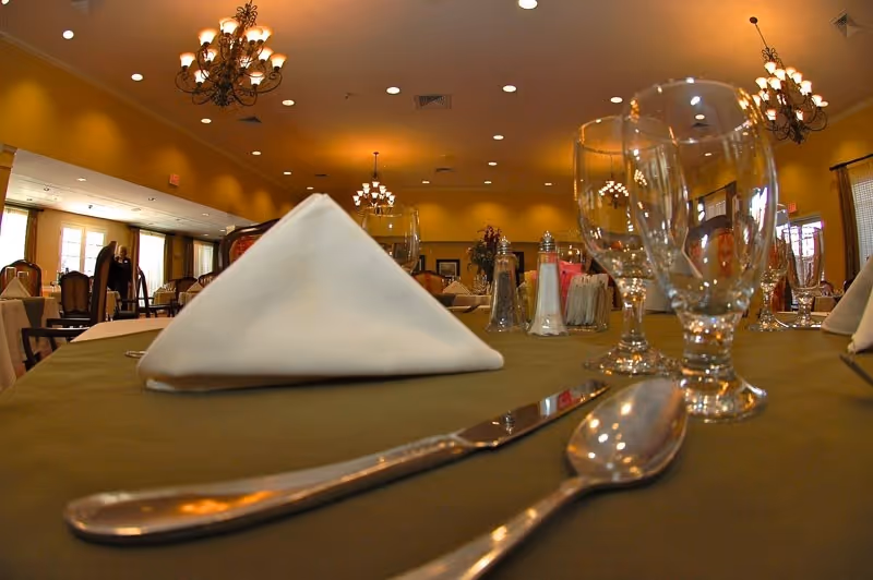 Close-up view of a dining table set with a folded white napkin, silverware, and empty glasses in a warmly lit dining room with chandeliers and multiple tables in the background.
