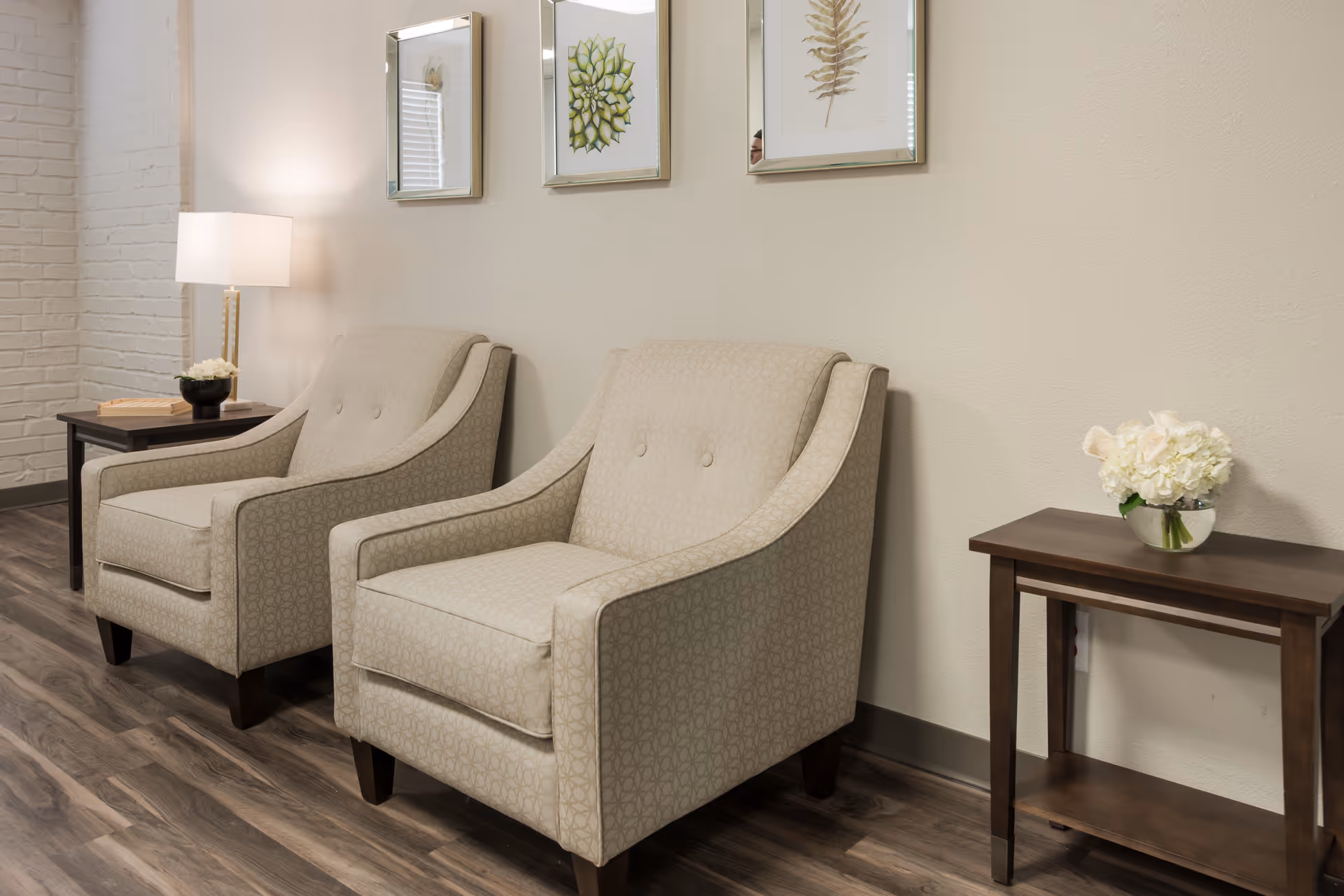 Two beige upholstered armchairs with side tables, a lamp, and framed wall art in a lounge-style seating area.